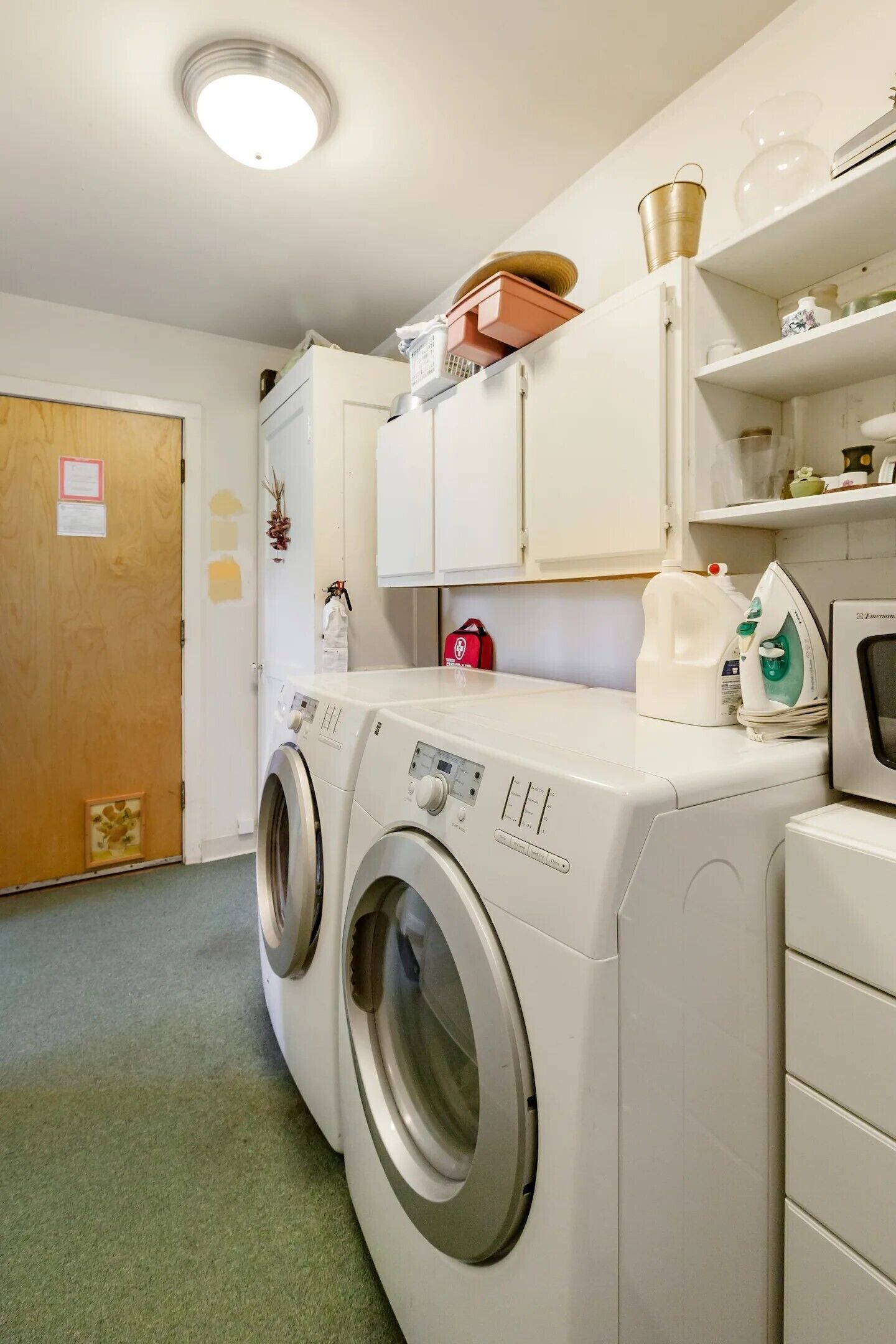 A laundry room with two front-loading washing machines, a dryer, and a shelf with various items.