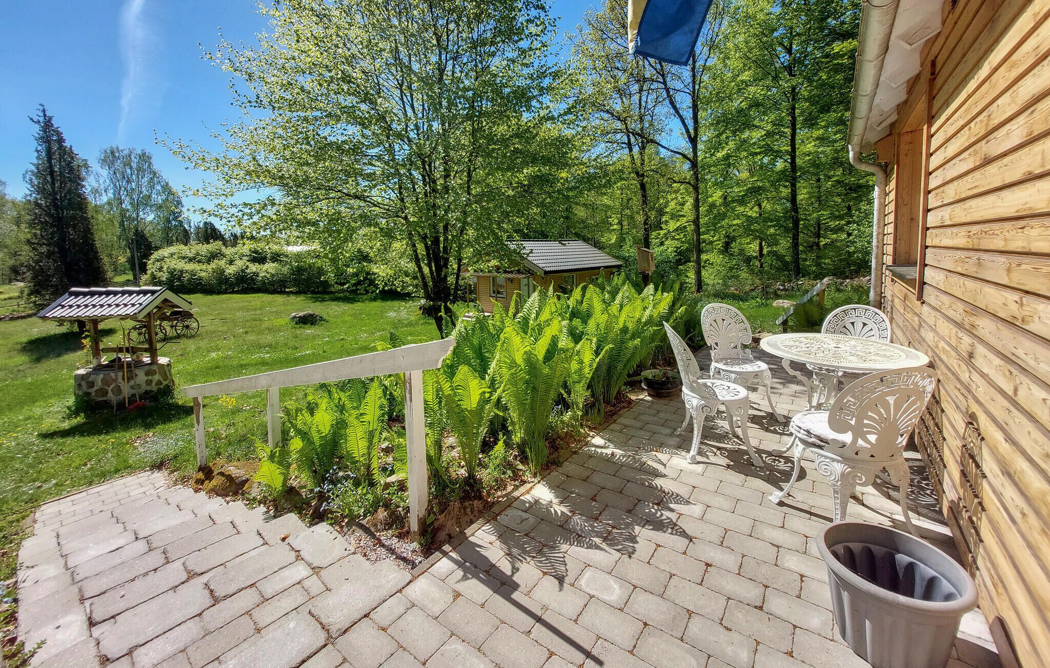 A patio area with a brick pathway leading to outdoor furniture, including a table and chairs, surrounded by lush greenery and a small building with a sloped roof.