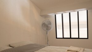Un dormitorio con cama, una ventana con persianas, un ventilador de pie y una toalla doblada sobre la cama.