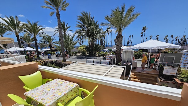 A terrace with a table and chairs, overlooking a street with palm trees and outdoor seating areas. - A009 Seaside view Las Vistas TRG Tenerife Royal Gardens (Playa de las Américas)