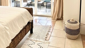 Une chambre d’hôtel comprenant un lit, un ventilateur de plafond, une télévision, une porte-fenêtre coulissante donnant sur un balcon aménagé avec du mobilier d’extérieur, et un panier en osier.