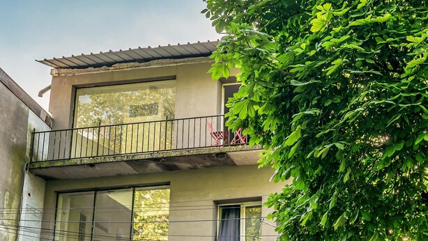 Un bâtiment à deux étages doté d’un balcon, de grandes fenêtres et d’un arbre au premier plan.