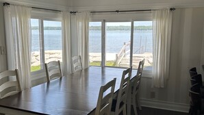 Ein Esszimmer mit einem großen Tisch und Stühlen, großen Fenstern mit weißen Vorhängen sowie Blick auf einen See und einen Strand.