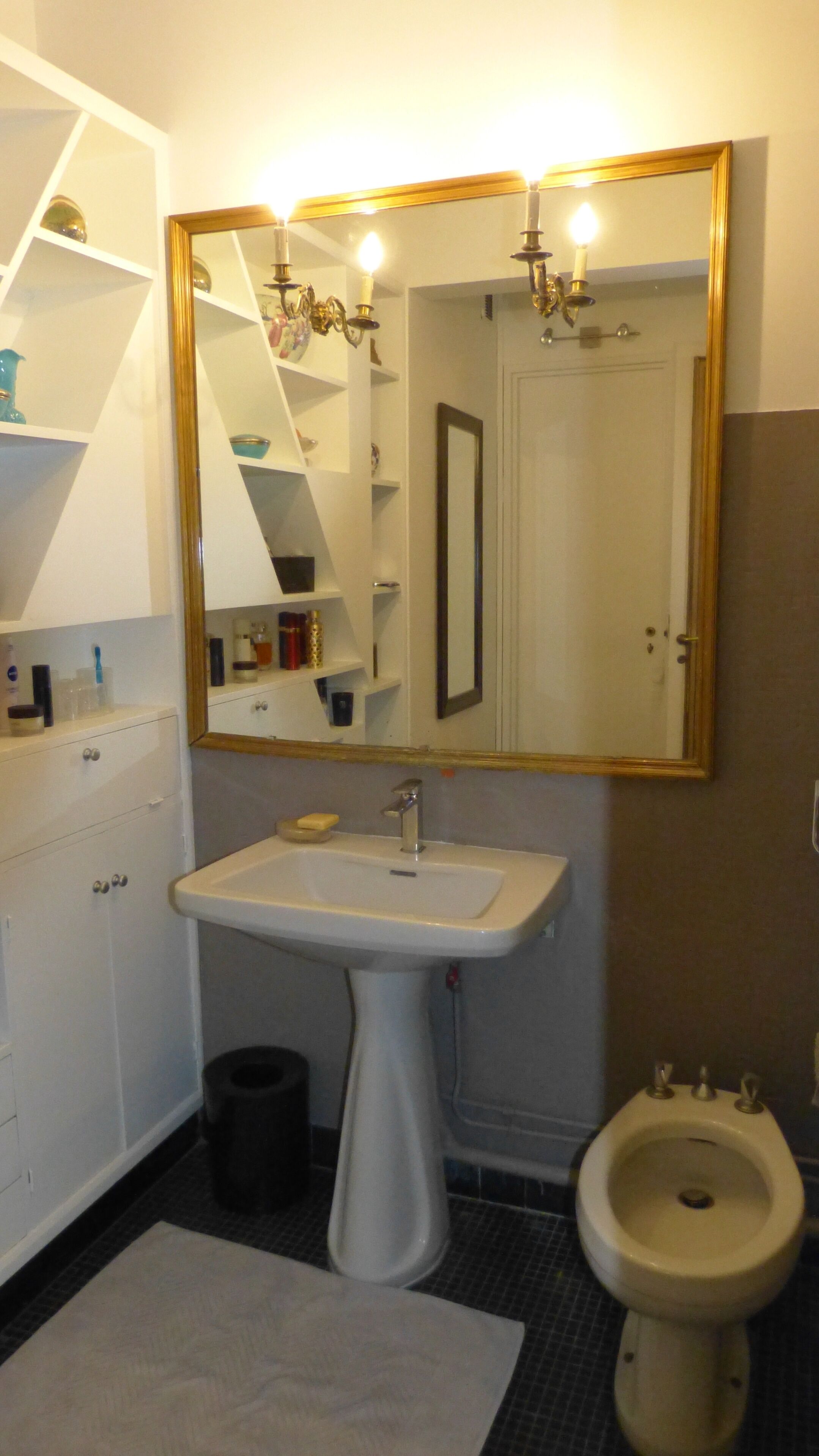 Ein Badezimmer mit einem Waschbecken auf Fuß, einem Bidet, einem Spiegel mit goldener Rahmung und einer weißen Kommode mit Regalen.