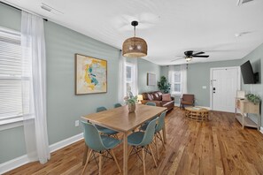 A spacious living area with a wooden dining table, teal chairs, a brown leather sofa, and a ceiling fan.