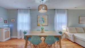Un comedor con una mesa de madera, cuatro sillas verdes, una lámpara colgante de fibras tejidas y un cuadro en la pared.