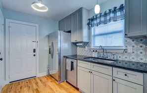 A kitchen with white cabinets, a stainless steel refrigerator, a black countertop, and a window with blinds.