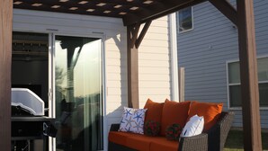 Une terrasse couverte avec un canapé en osier et des coussins, une porte vitrée et une pergola en bois.