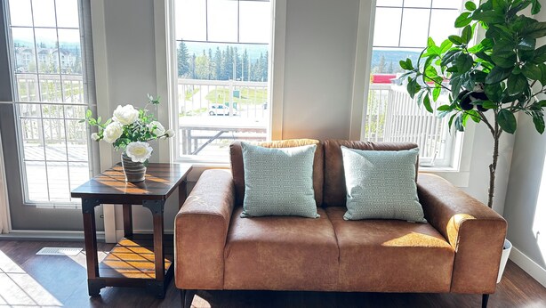 Ein braunes Ledersofa mit zwei gemusterten Kissen, ein kleiner Holztisch mit einer Vase weißer Blumen und eine große grüne Pflanze.
