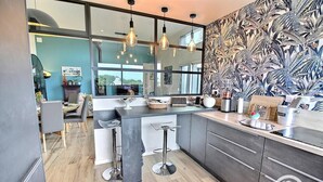 A modern kitchen with a large island, bar stools, and a view of the city through large windows. - Les Méloines - Trebeurden (TREBEURDEN)