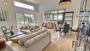A spacious living area with a large sofa, dining table, and chairs. There is a kitchen area with modern appliances and a large window providing a view of the outdoors. - Les Méloines - Trebeurden (TREBEURDEN)