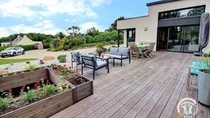 A wooden deck with outdoor furniture, a modern building with large windows, and a garden area with raised flower beds. - Les Méloines - Trebeurden (TREBEURDEN)
