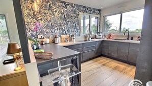 A modern kitchen with a dark backsplash, a central island, and a large window providing a view of the outdoors. - Les Méloines - Trebeurden (TREBEURDEN)