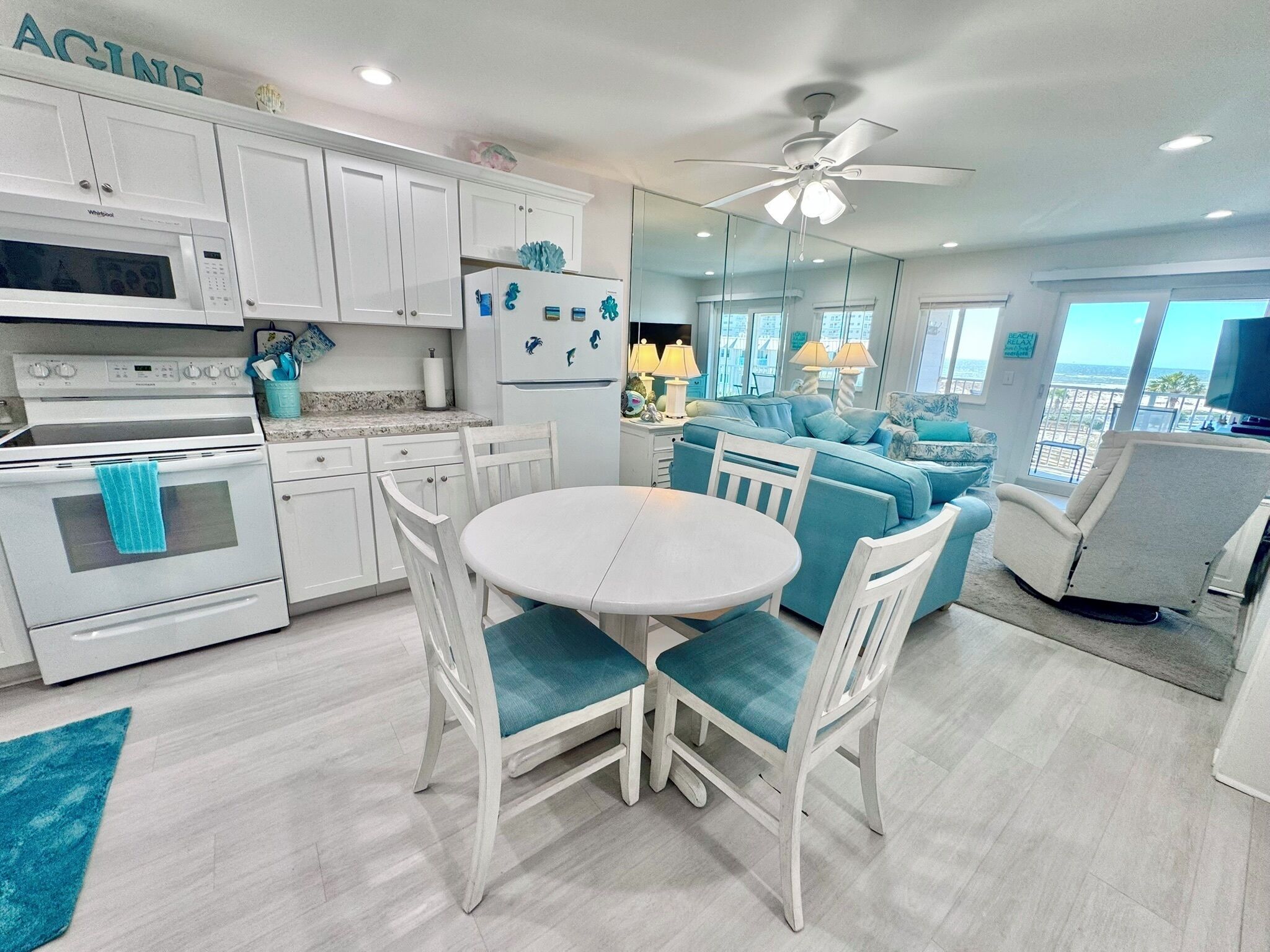 A modern kitchen with white cabinets, a white stove, and a dining area with a white table and blue chairs. - New Listing! Come check out the WAVES and catch a tan! (Gulf Shores)