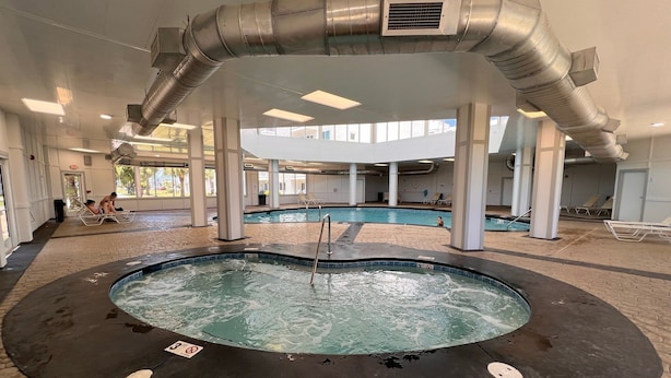 An indoor swimming pool with a hot tub, surrounded by lounge chairs and a covered area with a large air duct system.