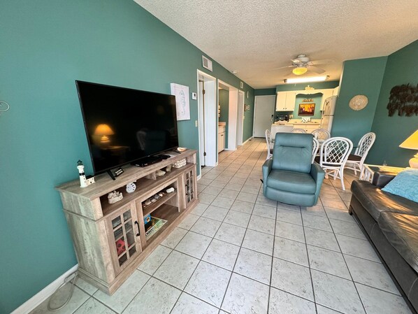 A living room with a TV on a wooden stand, a blue armchair, and a dining area with white chairs. - Ready for SUN, FUN and WAVES? Look no further! New Listing! (Gulf Shores)