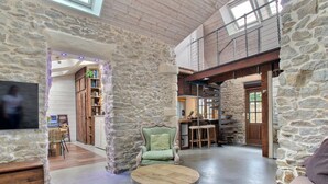 A spacious living area with stone walls, a television, a green armchair, and a kitchen in the background. - Echappée Breizh - Saint Pierre Quiberon (SAINT PIERRE QUIBERON)