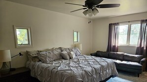 Un dormitorio con una cama, ventilador de techo, ventana con cortinas, mesita de noche con lámpara y un sofá.
