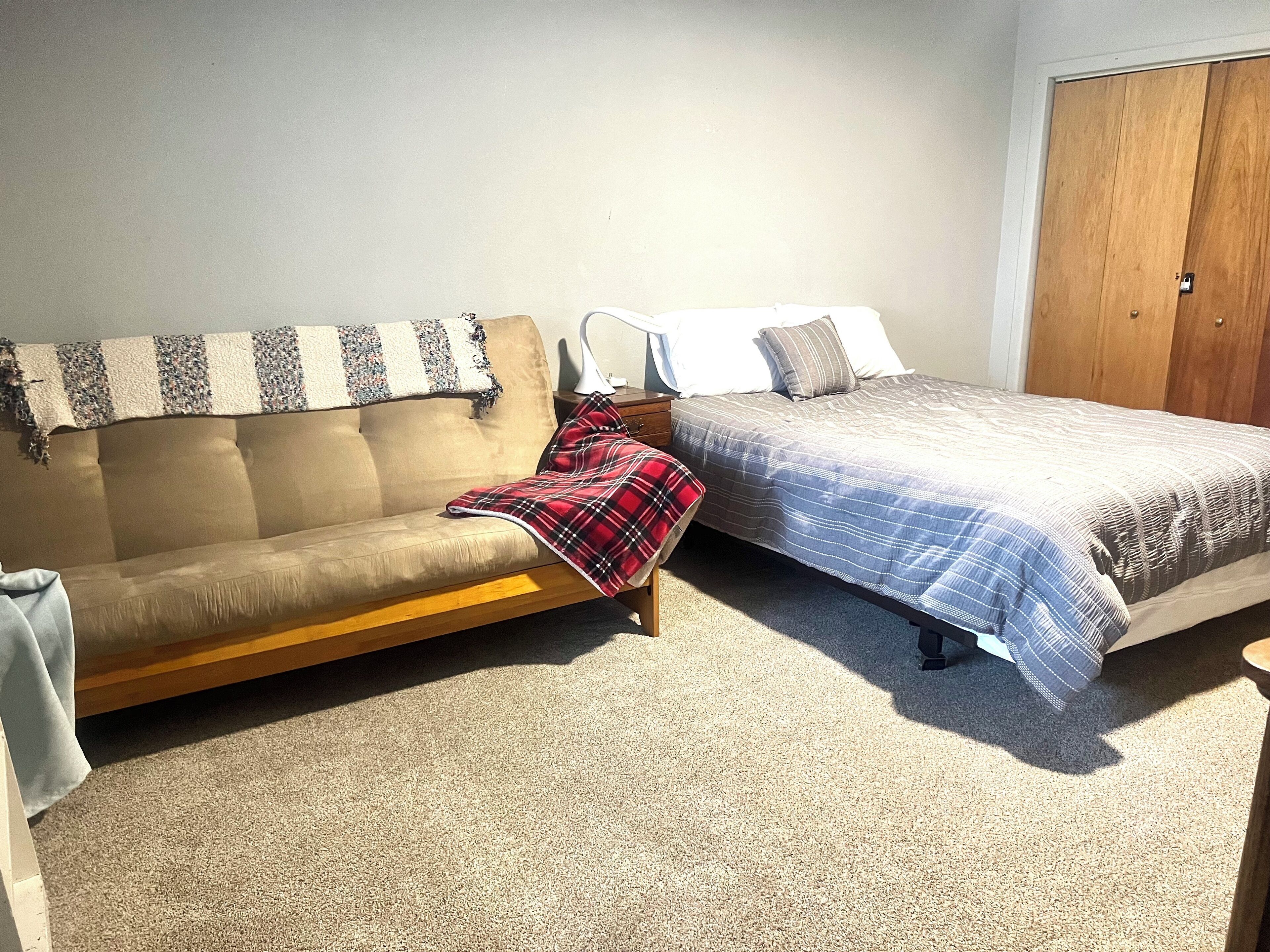 Een slaapkamer met een beige bank, een bed met een blauw-witte gestreepte dekbedovertrek en een houten garderobe.