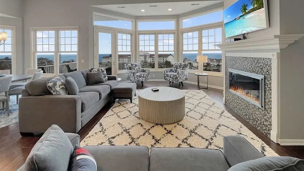 A spacious living room with a large gray sectional sofa, a round coffee table, and a fireplace. The room features large windows offering a view of the cityscape.