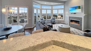 A spacious living room with a fireplace, a large TV, and a view of the ocean through large windows.