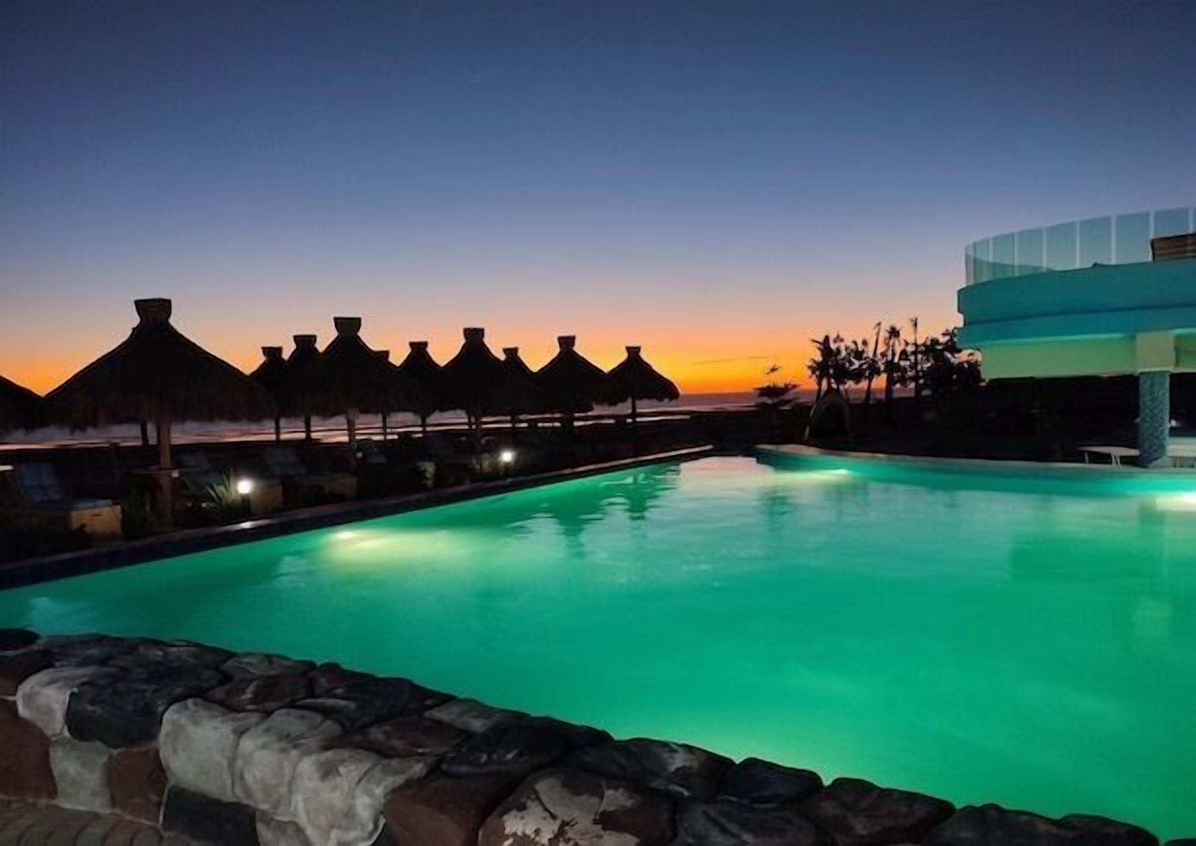Une piscine au crépuscule, avec des parasols en chaume et un bâtiment moderne en arrière-plan.