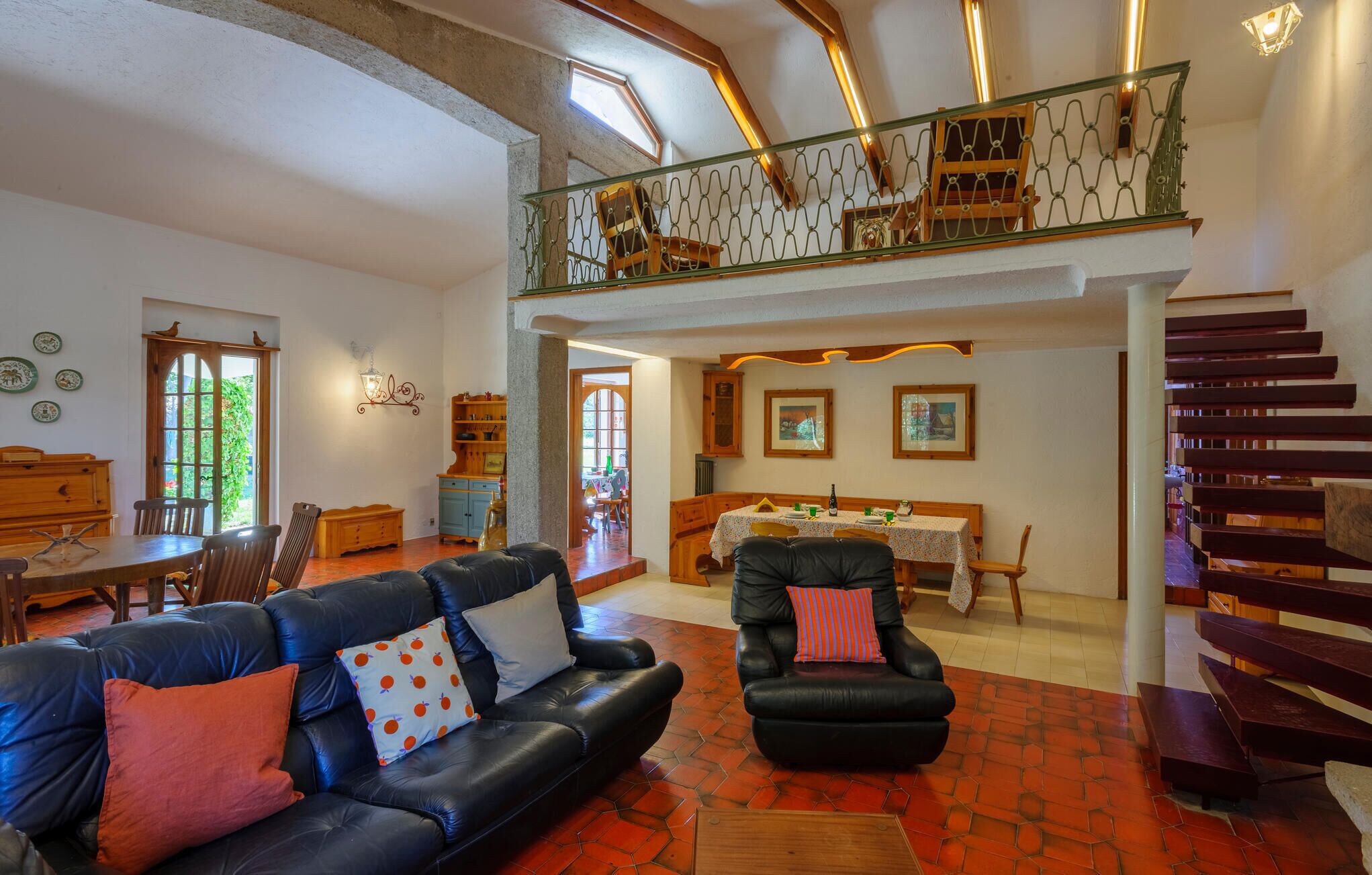 Un salón espacioso con un sofá de cuero negro, una mesa de comedor de madera y una escalera que sube a un nivel superior.