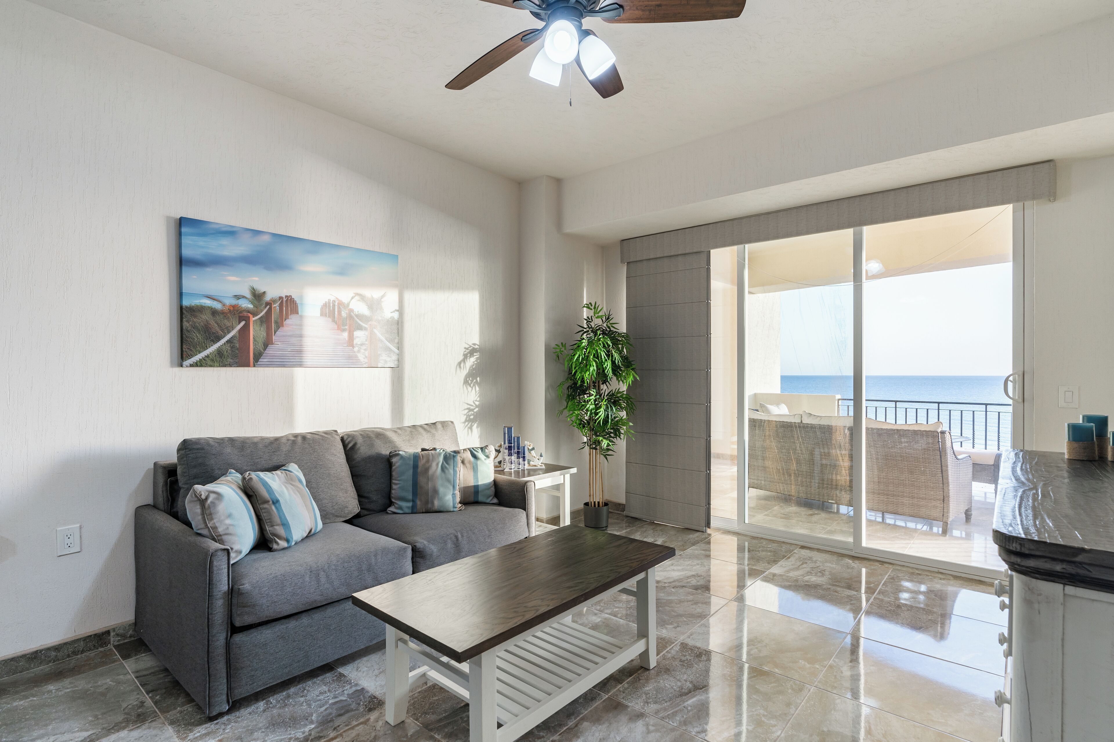 A modern living room with a grey sofa, a wooden coffee table, and a large window overlooking the ocean.