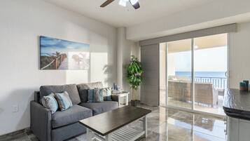 A modern living room with a grey sofa, a wooden coffee table, and a large window overlooking the ocean.