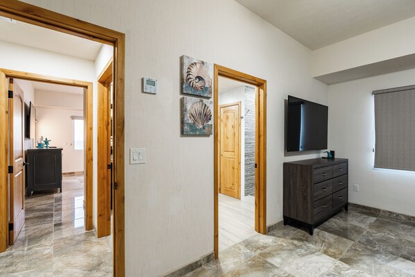 A hallway with a wooden door, a dark wooden dresser, a flat-screen TV mounted on the wall, and a window with a blind.