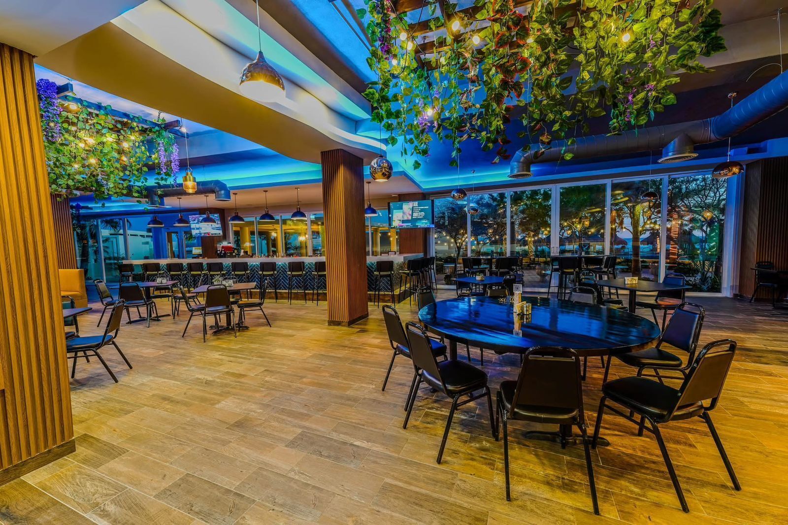 A spacious indoor dining area with multiple tables and chairs, a wooden floor, and a ceiling adorned with hanging plants and lights.