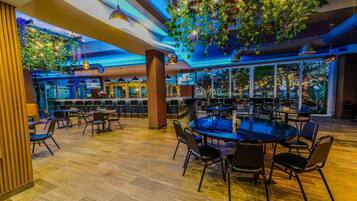 A spacious indoor dining area with multiple tables and chairs, a wooden floor, and a ceiling adorned with hanging plants and lights.
