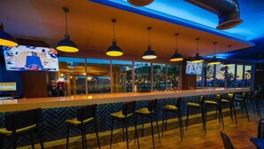 Een bar met een lange toonbank, meerdere hangende lampen en een groot televisiescherm waarop een sportwedstrijd te zien is.