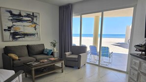 A living room with a sofa, armchair, and coffee table, a painting on the wall, and a view of the beach through large glass doors.