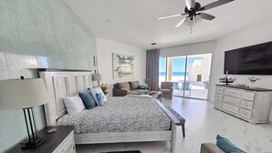 A spacious bedroom with a large bed, a ceiling fan, and a view of the sea through large windows.