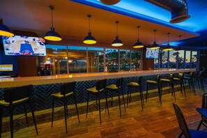 Un espace bar doté d’un long comptoir, de plusieurs luminaires suspendus et d’un écran de télévision diffusant un événement sportif.