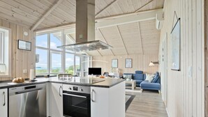 A modern kitchen with a built-in oven and dishwasher, large windows offering a view of the outdoors, and a cozy living area with a blue sofa and a flat-screen TV. - 3 bedroom awesome home in Faaborg (Faaborg)