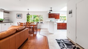 A modern kitchen with wooden cabinets, a dining area with a brown leather sofa, and a view of greenery through the window. - Coastal ranch-style home in garden setting.  (Carpinteria)