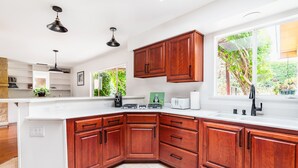A modern kitchen with wooden cabinets, a white countertop, and a large window offering a view of greenery outside. - Coastal ranch-style home in garden setting.  (Carpinteria)