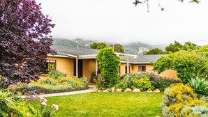 A yellow house with a garden in front, surrounded by mountains and fog. - Coastal ranch-style home in garden setting.  (Carpinteria)