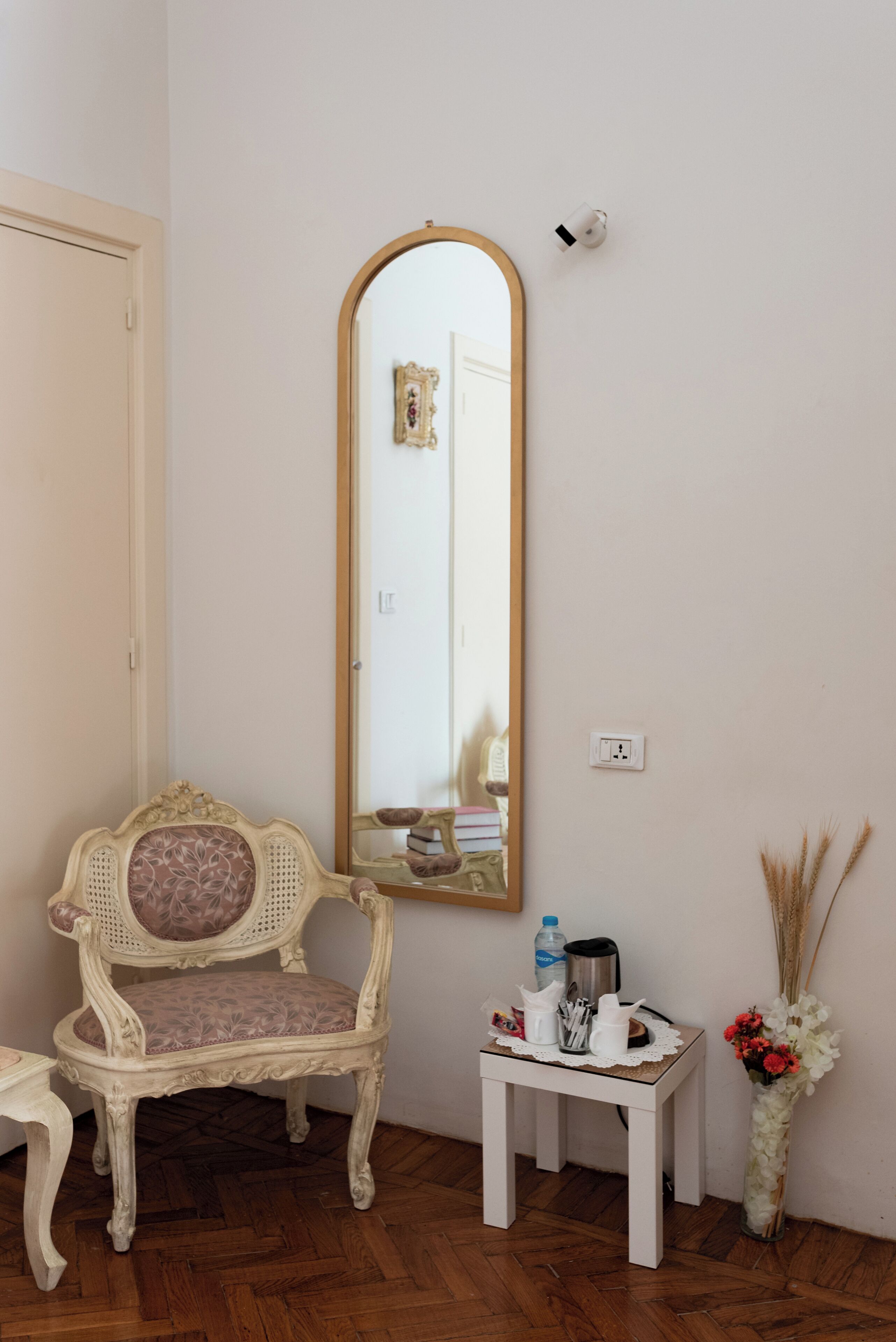 A room with a wooden floor, a white chair with a floral cushion, a small white table with a vase of dried plants, and a tall mirror on the wall.