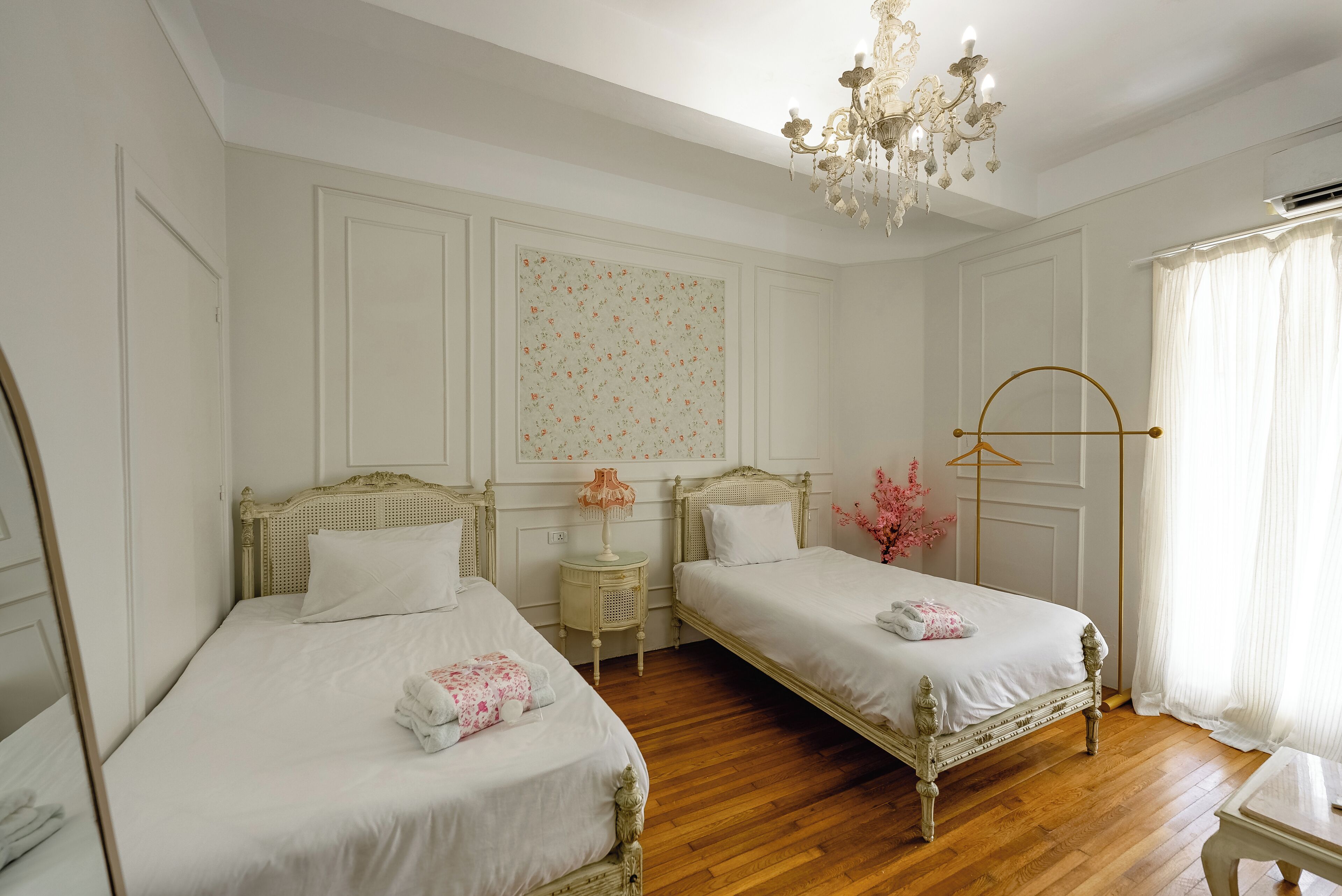 Quarto de hotel com duas camas de solteiro, um candelabro, um espelho e uma janela com cortinas.