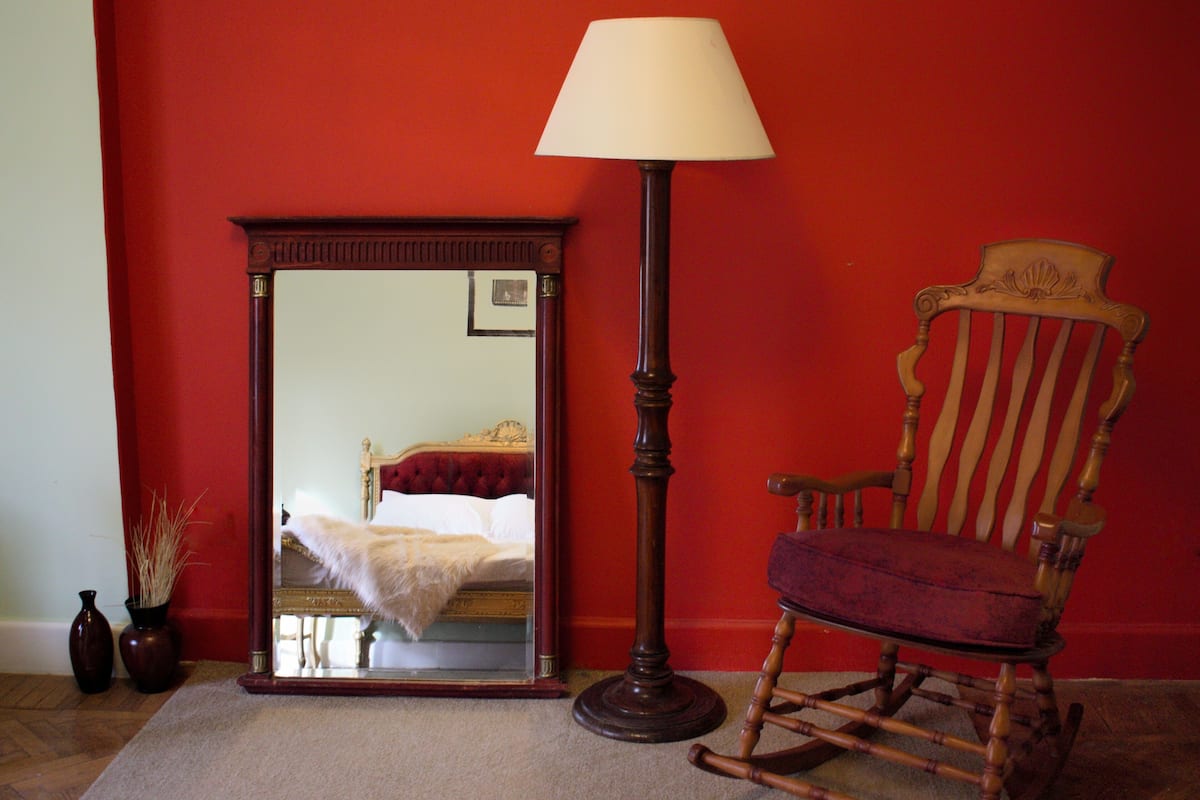 A room with a mirror, a rocking chair, and a floor lamp against a red wall.