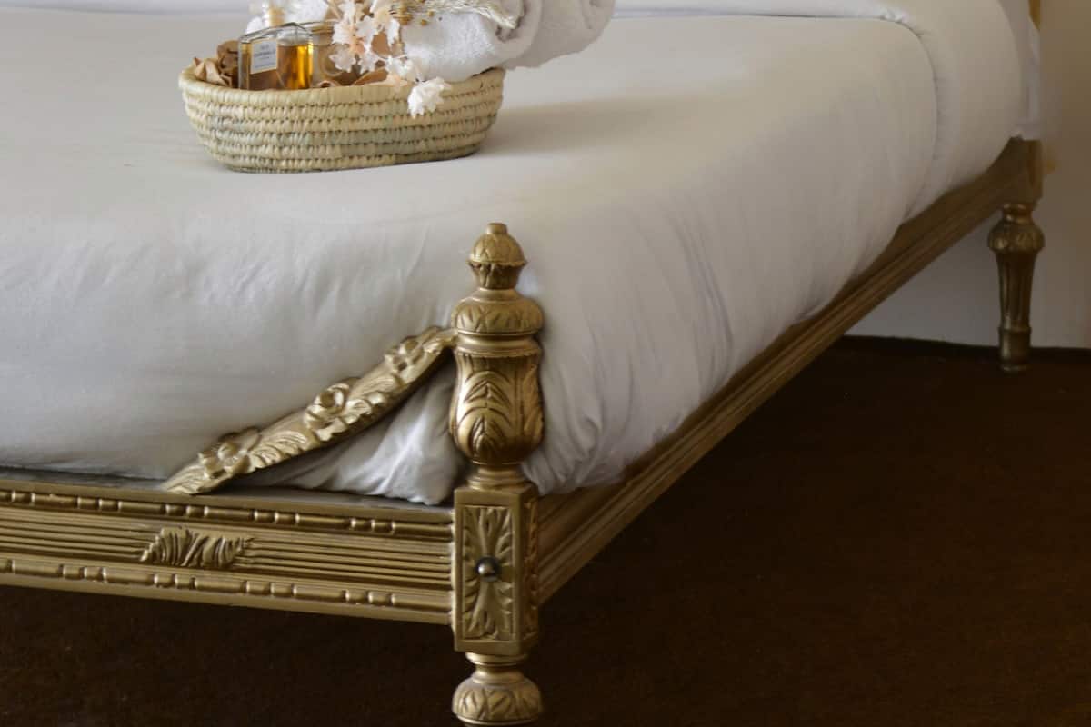A bed with a woven headboard, white linens, a brown pillow, and a basket with flowers and a bottle.