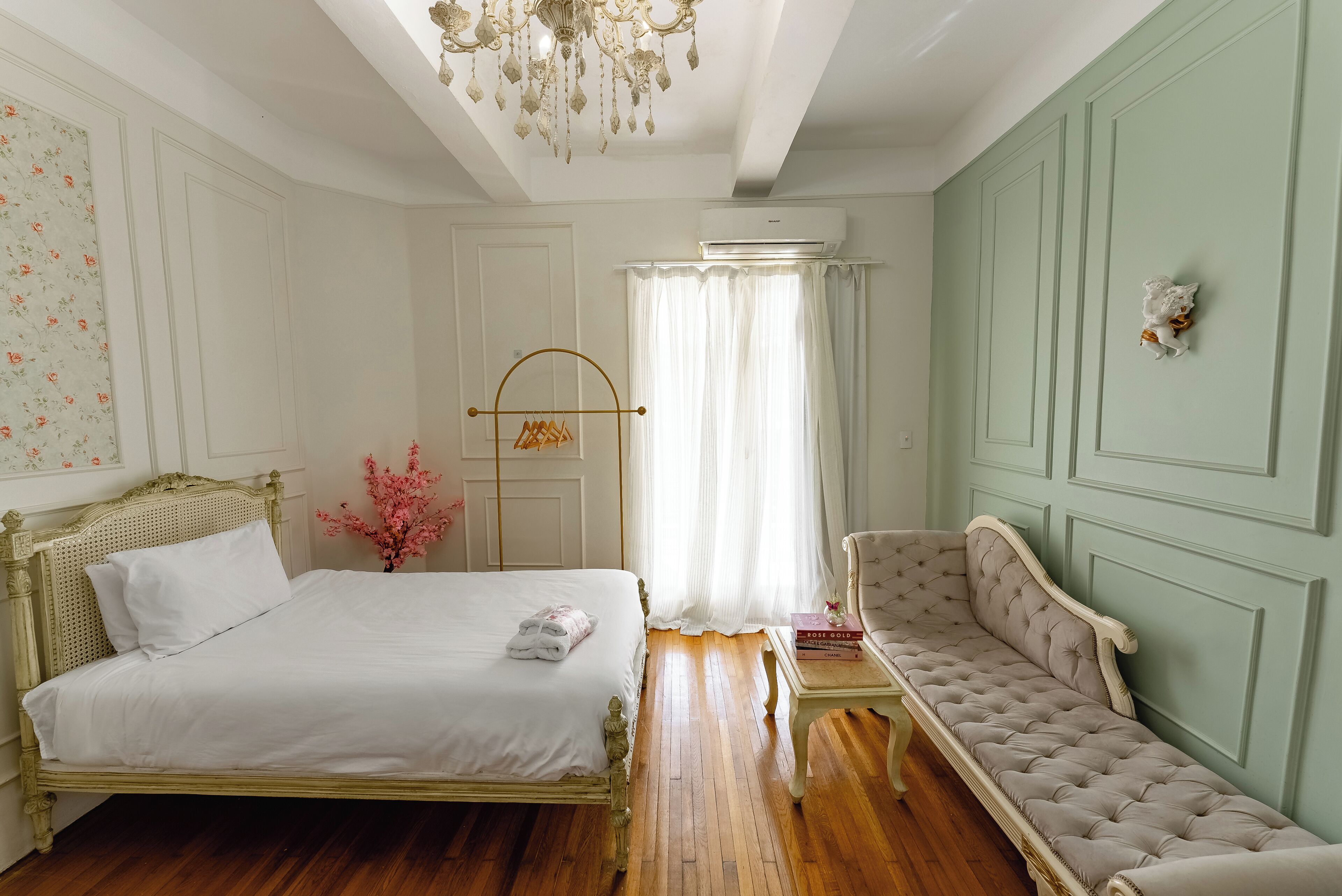 A bedroom with a bed, a bench, a chandelier, and a window with curtains.