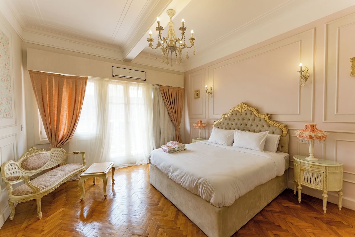 A bedroom with a large bed, a chaise lounge, a small table, and a chandelier.