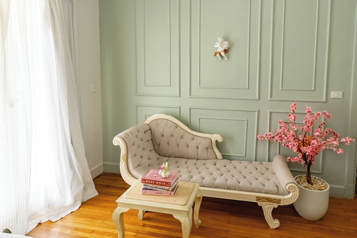 A room with a light green wall, a tufted sofa, a small table with books, and a potted plant with pink flowers.