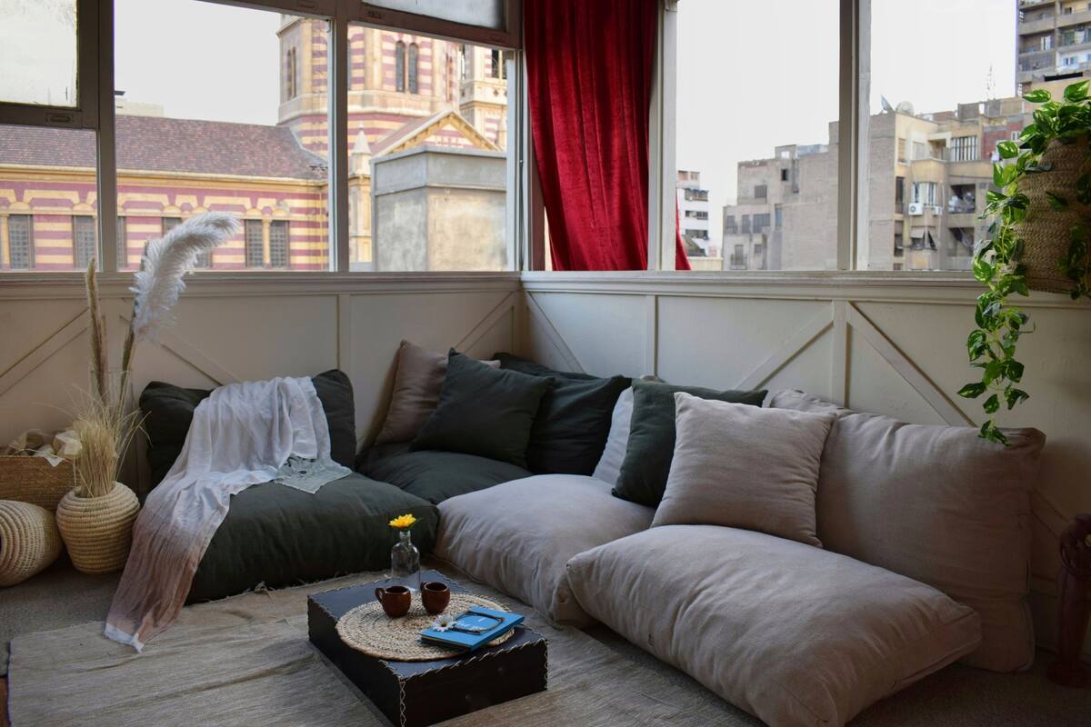 A cozy living room with a large window, a sofa, a small table with a book and two cups, and a decorative feather.