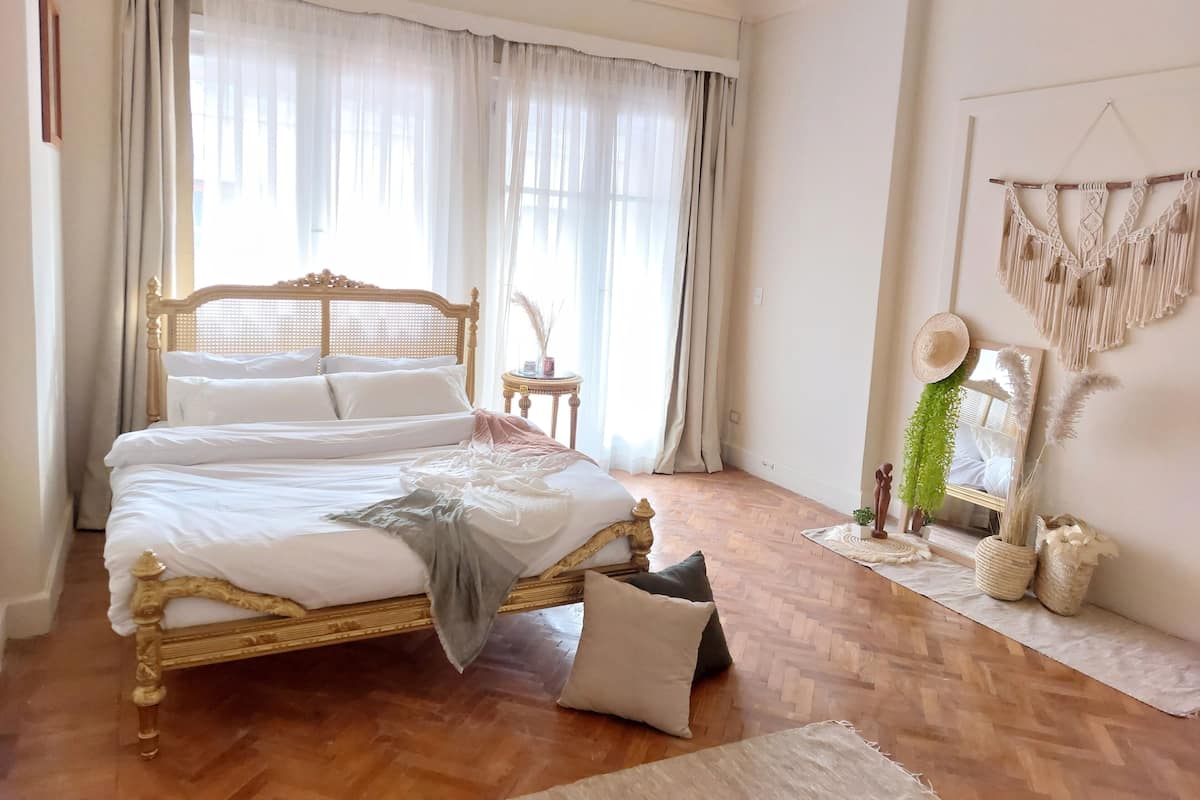 A bedroom with a wooden bed, white bedding, a small round table, and a macrame wall hanging.