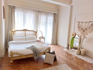 Ein Schlafzimmer mit einem hölzernen Bett, weißer Bettwäsche, einem kleinen runden Tisch und einem Wandbehang aus Makramee.